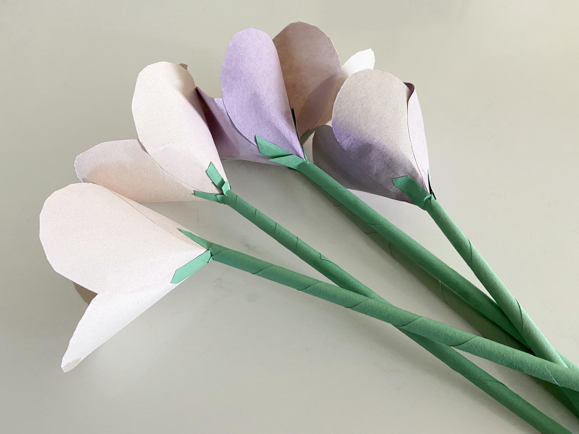 A faded bouquet of paper flowers lays on a white counter top. There are two pink and three purple tulip-like flowers made of construction paper. Their stems are drinking straws wrapped in green construction paper.